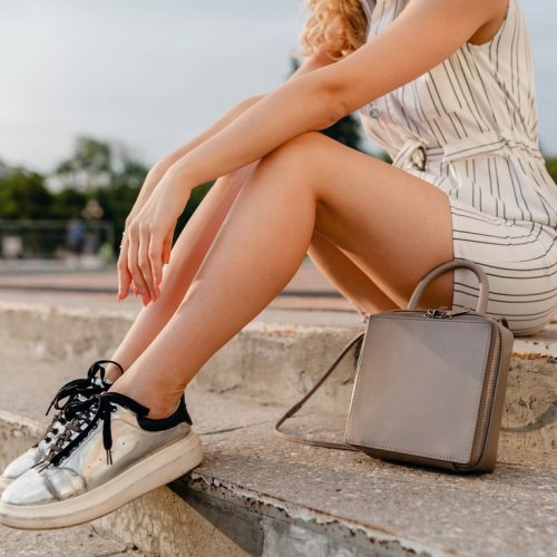 accessories of stylish woman walking in city street in summer fashion style, legs in sneakers, grey purse handbag