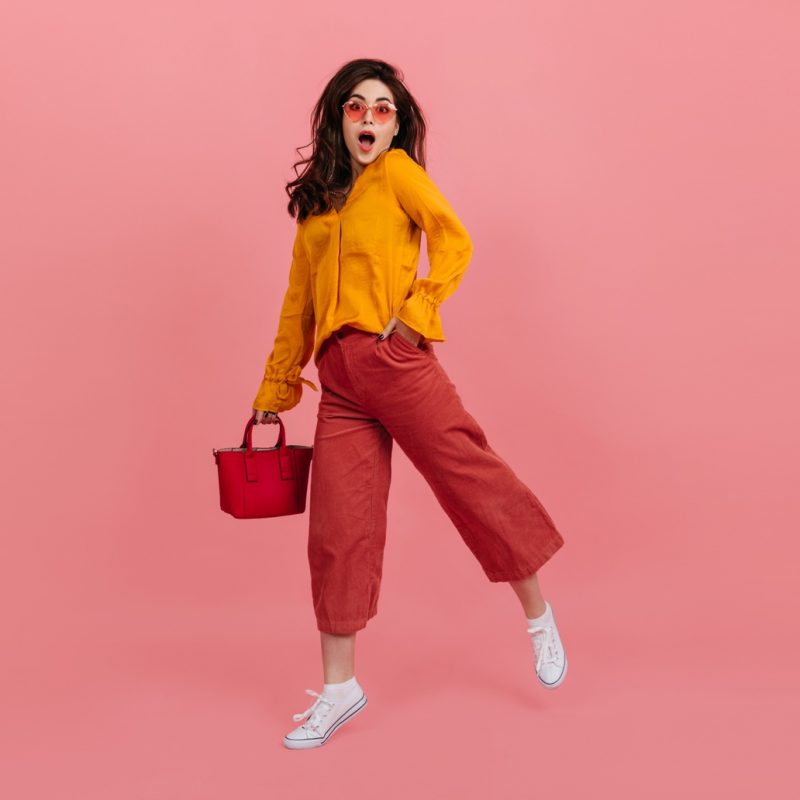 Perky girl in stylish glasses stares into camera in amazement, walking on pink background. Brunette in culottes and orange blouse posing with red handbag